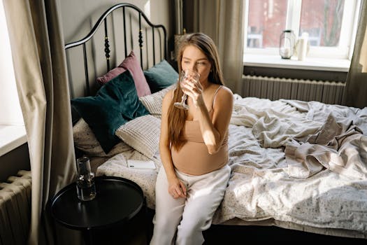 Pregnant woman sitting on bed, drinking water in a cozy bedroom setting.